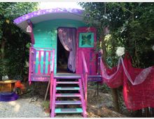 Piccola casetta in legno color turchese e fucsia con decorazioni floreali, scaletta e veranda con ringhiere, immersa tra alberi e vegetazione. L’ingresso ha una tenda viola e accanto si vede un drappo di tessuto rosso appeso tra i rami.