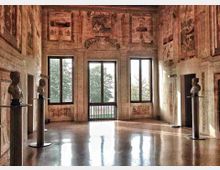 Sala storica con pareti affrescate e soffitto alto, illuminata da grandi finestre che affacciano su alberi all’esterno. Pavimento in marmo lucidato e busti scultorei su piedistalli disposti lungo i lati della stanza.