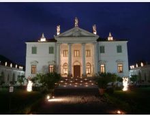 Facciata notturna di una villa neoclassica simmetrica, con portico centrale a colonne e frontone triangolare sormontato da statue. Una scalinata illuminata conduce all’ingresso, con giardino e luci decorative ai lati.