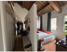 Interno di una camera con soffitto alto a travi di legno e pavimento in legno, divisa da una parete: da un lato armadi e panca lungo il corridoio, dall’altro una zona letto con finestra e tende scure. Arredi moderni essenziali e luce naturale proveniente dalle finestre.
