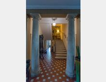 Ingresso interno con due grandi colonne in pietra che incorniciano un corridoio con pavimento a piastrelle rosse e motivi geometrici. In fondo si vede una scala in pietra che sale verso una parete con carta da parati decorata, illuminata da lampade a sospensione.