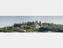 Panorama di una collina verde con alberi alti e cipressi, e diversi edifici storici in pietra tra cui una torre e una chiesa. In primo piano si vedono terrazzamenti e muri di contenimento, sotto un cielo limpido.