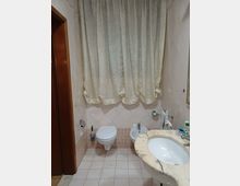 Bagno domestico con pareti e pavimento piastrellati in tonalità beige, wc sospeso e bidet affiancati sotto una grande finestra coperta da tende chiare. In primo piano si vede un lavabo incassato in un piano in marmo, con porta in legno sul lato sinistro.