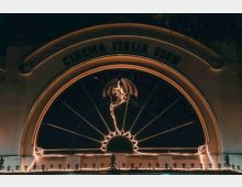 Insegna ad arco illuminata di un cinema all’aperto, con la scritta “CINEMA ITALIA EDEN” e un fregio centrale a forma di drago. Sul bordo inferiore si vede una ringhiera decorativa con luci lungo il profilo.