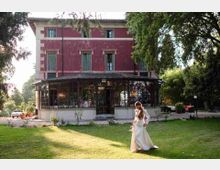 Villa storica a tre piani con facciata rosso scuro e finestre alte, con una veranda chiusa in vetro e metallo al piano terra. Davanti si estende un prato verde con alberi e vegetazione ai lati.