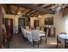 Sala da pranzo in stile rustico con soffitto a travi di legno a vista e lampadari in vetro, con un lungo tavolo centrale coperto da tovaglia floreale e sedie imbottite. Lungo le pareti ci sono credenze e armadi in legno scuro, quadri appesi e una grande finestra laterale con tende che illumina l’ambiente.