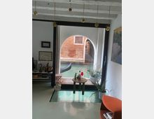 Interno di uno spazio moderno con pavimento lucido, decorato con una poltrona arancione, un tavolino e piante, attraverso una grande finestra ad arco che dà su un canale veneziano con una gondola visibile sull'acqua. Il soffitto presenta travi in legno bianche.