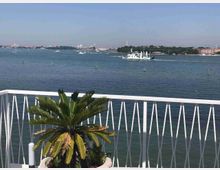 Terrazza con ringhiera bianca e una pianta in vaso in primo piano, affacciata su un’ampia laguna. Sullo sfondo si vedono imbarcazioni in navigazione e la linea costiera con edifici e campanili.