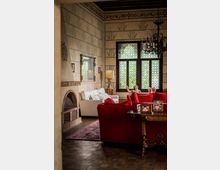 Soggiorno in stile classico con pareti decorate a motivi geometrici, camino in muratura e grande finestra a tre ante con vetri a rosoni. Arredi con divani in tessuto rosso e bianco, tappeto orientale, tavolino in legno e lampadari in ferro battuto.