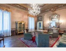 Ampio salone storico con soffitto affrescato e riccamente decorato, grande lampadario in cristallo e pavimento in parquet. Arredi classici con poltrone attorno a un tavolo su tappeto, vetrina e grande specchio dorato, e alte finestre con tende chiare.