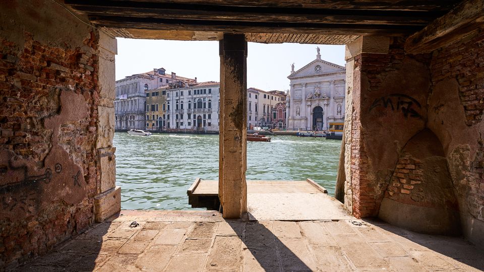 Vista da un portico in mattoni e pietra con travi in legno affacciato su un canale, con piccola banchina sull’acqua. Sullo sfondo si vedono palazzi storici lungo la riva e una chiesa con facciata classica in pietra chiara.