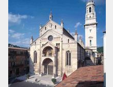Facciata di una grande chiesa romanica in pietra chiara con portale ad arco, rosone centrale e finestroni laterali, affacciata su una piazza. Sul lato destro si innalza un alto campanile bianco a più livelli, con tetti e edifici storici circostanti sotto un cielo azzurro.