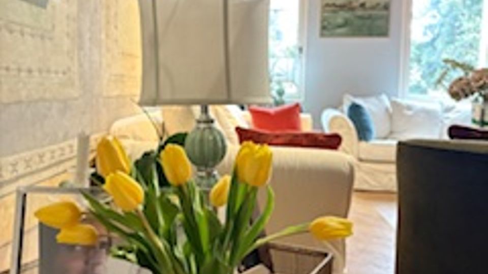 Interno di un soggiorno luminoso con pavimento in parquet a spina di pesce e travi in legno a vista. In primo piano un tavolino metallico con lampada e vaso di tulipani gialli, sullo sfondo un divano chiaro vicino a grandi finestre.