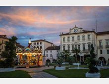 Piazza cittadina al tramonto con aiuole e vialetti in pietra; al centro è presente una giostra a cavalli illuminata. Sullo sfondo si vede un edificio storico con facciata chiara, finestre ad arco, porticato e un orologio sulla sommità.