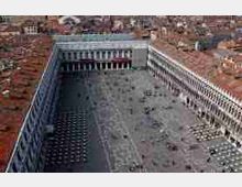 Vista aerea di una grande piazza rettangolare lastricata, circondata su tre lati da lunghi edifici porticati con arcate regolari e facciate in pietra. Sullo sfondo si vede un’area urbana fitta con tetti in tegole rosse.