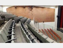 Interno di un auditorium con file di sedute grigie disposte a gradinate e corridoi laterali. In fondo si vede una grande parete in mattoni, una balconata con parapetto in vetro e, sul lato destro, il bordo del palco con tendaggi rossi.