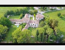 Veduta aerea di un grande complesso rurale in stile cascina, con edifici a tetto in coppi disposti attorno a un cortile interno e circondati da giardini alberati. Intorno si vedono campi verdi, viali e una strada di campagna con incrocio nelle vicinanze.