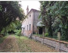 Edificio storico in mattoni rossi con piccole finestre circolari e coperture ad arco sugli ingressi, affacciato su un sentiero sterrato. L’area è circondata da vegetazione fitta e alberi, con un basso muretto in mattoni lungo il lato destro.