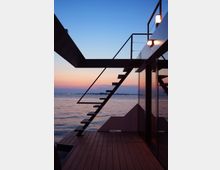 Terrazza moderna in legno affacciata su un ampio specchio d’acqua al tramonto, con struttura in metallo e scale sospese che conducono a un livello superiore. Pareti vetrate e corrimano minimalisti incorniciano il cielo sfumato e l’orizzonte sull’acqua.