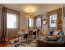 Sala da pranzo in un appartamento, con tavolo rettangolare e sedie imbottite sotto un lampadario classico. Grandi finestre con tende leggere, pavimento in legno con tappeto, divano laterale e specchio rotondo sulla parete.