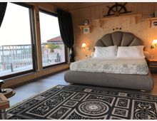 Camera da letto con pareti in legno e letto matrimoniale con testiera imbottita, illuminato da applique e mensole decorative. Grandi porte-finestre ad angolo con tende scure si affacciano su un terrazzo con ringhiera e vista sul mare.