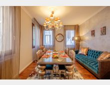 Sala da pranzo e soggiorno in un ambiente unico, con tavolo rettangolare apparecchiato e sedie imbottite al centro, divano blu trapuntato lungo la parete e pavimento in legno con tappeto. Grandi finestre con tende leggere, lampadario classico a più luci, specchio rotondo a parete e lampada da terra completano l’arredo.