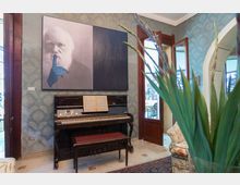 Salotto interno con carta da parati decorata, pianoforte verticale con spartito e panca, e grande quadro moderno appeso alla parete. Porte finestre in legno aperte su una terrazza esterna e arco di passaggio sul lato destro.