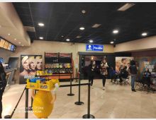 Atrio interno di un cinema con pavimento chiaro e controsoffitto nero illuminato da faretti, banco snack a sinistra e scaffali di caramelle al centro. Sullo sfondo si vedono locandine di film, un’insegna “Paradise” sopra un ingresso e una zona con tavolini e sedie sulla destra.