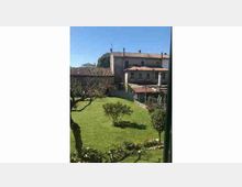 Vista su un giardino con prato curato e alberi, circondato da edifici in stile tradizionale con facciate chiare e tetti in tegole. Sullo sfondo si vedono muri di recinzione e una piccola struttura coperta all’interno del cortile.