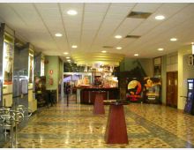 Atrio interno di un cinema con pavimento a mosaico, soffitto a pannelli con luci incassate e un banco bar centrale sotto una pensilina circolare. Lungo le pareti si vedono locandine e ingressi alle sale, con una scala che sale sul lato destro.