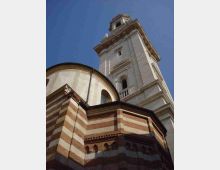 Vista dal basso di una chiesa storica con facciate a fasce orizzontali in mattoni e pietra e un’abside semicircolare. Accanto si innalza un alto campanile in pietra chiara con finestre ad arco e cornici modanate, sullo sfondo di cielo azzurro.