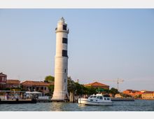 Faro cilindrico in pietra chiara con una fascia nera e balconata in cima, situato su una banchina lungo l’acqua. In primo piano si vede un canale con una barca, mentre sullo sfondo compaiono edifici con tetti in tegole rosse e un cantiere con gru.
