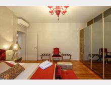 Camera da letto moderna con pavimento in parquet, letto con tessili rossi e lampada da comodino decorativa. Di fronte ci sono una poltrona imbottita rossa, due panche portavaligie e un grande armadio a specchio, con lampadario rosso a soffitto e climatizzatore sopra la porta.