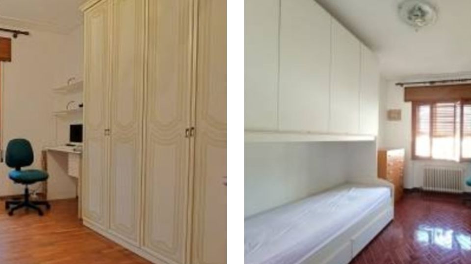 Interno di una camera da letto luminosa con pavimento in legno, armadiature alte a tutta parete, letto e piccola scrivania con sedia. Una grande finestra con persiane in legno e termosifone sottostante illumina l’ambiente.