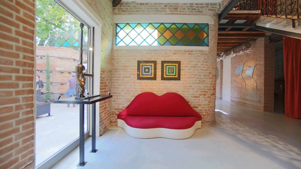 Interno di uno spazio espositivo in stile industriale con pareti in mattoni a vista, pavimento chiaro e travi in legno, illuminato da una grande porta vetrata. Al centro c’è un divano rosso a forma di labbra sotto una finestra con vetri colorati, con quadri alle pareti e un soppalco con ringhiera in ferro battuto.