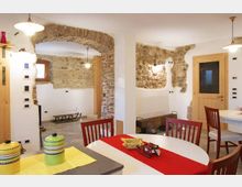 Interno di una cucina-sala da pranzo con pareti bianche e inserti in pietra a vista, arco in muratura e pavimento in piastrelle. Tavolo con runner rosso e sedie in legno, piano cucina con accessori, porta in legno e lampade a sospensione.