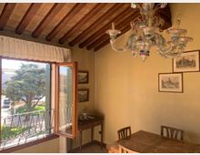 Interno di una sala da pranzo in stile rustico con soffitto a travi di legno a vista, lampadario in vetro e tavolo con sedie in legno. Una grande finestra aperta con ringhiera in ferro battuto affaccia su un’area esterna alberata e illuminata dal sole, con quadri appesi alle pareti.