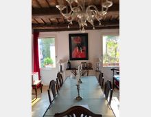 Sala da pranzo in stile rustico con lungo tavolo rettangolare e sedie scure, lampadario decorativo e soffitto con travi in legno a vista. Due finestre con tende rosse illuminano la stanza, con credenza sul fondo e un quadro appeso alla parete.