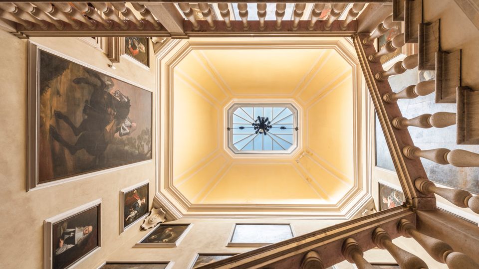 Veduta dal basso di un vano scala interno con ringhiere in pietra e pareti decorate da grandi dipinti incorniciati. Al centro si apre un lucernario ottagonale con vetrata e lampadario, incorniciato da un soffitto a riquadri color crema.