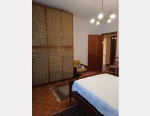 Camera da letto con pavimento in parquet, grande armadio a tutta parete con ante rivestite a motivo geometrico e letto in legno con copriletto bianco. A destra si vedono una porta aperta verso il corridoio, una sedia in legno e un lampadario a soffitto con più luci.