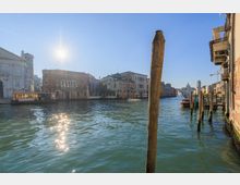 Veduta di un ampio canale urbano con acqua verde e riflessi del sole, fiancheggiato da palazzi storici e una chiesa sulla sinistra. In primo piano si vede un palo d’ormeggio e, sulla destra, una fila di pali e piccole imbarcazioni lungo la riva.