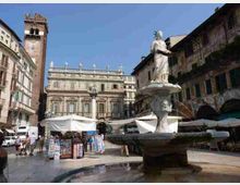 Piazza cittadina con una fontana in pietra e statua al centro, circondata da bancarelle e tende bianche. Sullo sfondo si vedono edifici storici con facciate ornate e una torre campanaria, sotto un cielo sereno.