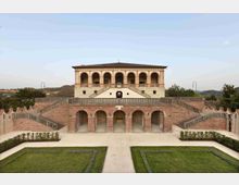 Facciata di una villa storica sopraelevata con loggiato ad archi, grande terrazza centrale e doppia scalinata simmetrica in muratura. In primo piano giardino geometrico con aiuole e vialetti, sullo sfondo colline e cielo sereno.