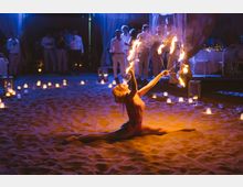 Spazio eventi su una spiaggia sabbiosa di notte, illuminato da numerose candele e lanterne disposte a terra. Sullo sfondo si vedono tende e tavoli apparecchiati con luci soffuse in un’area all’aperto.