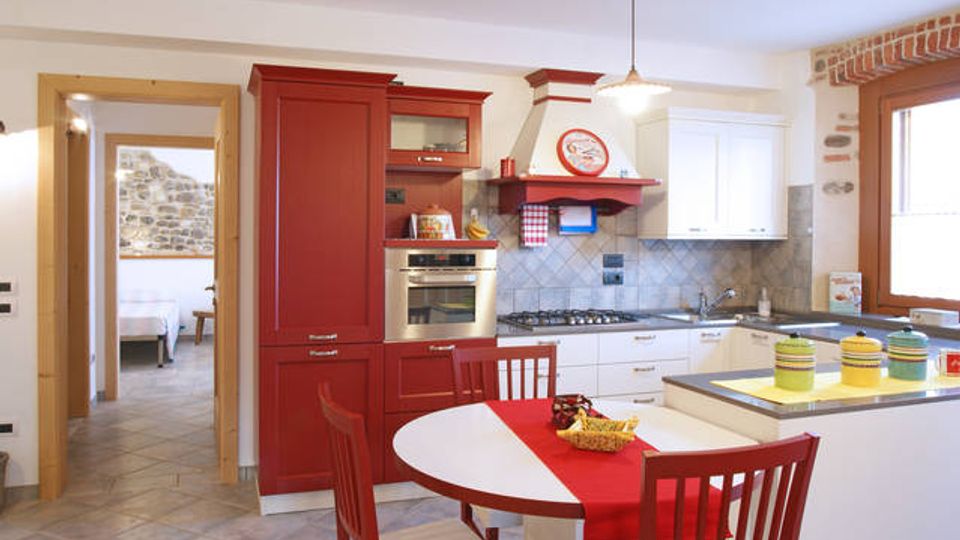 Cucina abitabile in stile rustico con mobili bianchi e rossi, forno incassato, piano cottura e cappa con mensola. In primo piano un tavolo rotondo con sedie rosse e runner, e sullo sfondo una porta verso un’altra stanza con parete in pietra e una finestra che illumina l’ambiente.