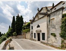 Piccola chiesa storica intonacata bianca lungo una strada curva, con facciata a timpano decorata da statue e una finestra trilobata sopra l’ingresso. Accanto ci sono muri in pietra coperti di vegetazione e alti cipressi, con cielo parzialmente nuvoloso sullo sfondo.