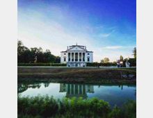 Villa in stile neoclassico con facciata bianca e alto portico con colonne, affacciata su un canale che ne riflette l’immagine. Davanti si vede un argine erboso e, ai lati, alberi e vegetazione lungo il percorso.