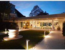 Esterno serale di un edificio moderno a un piano con ampie vetrate che mostrano un salone illuminato all’interno. In primo piano ci sono un giardino con prato, una fontana decorativa e un vialetto pavimentato con piccoli lampioni.