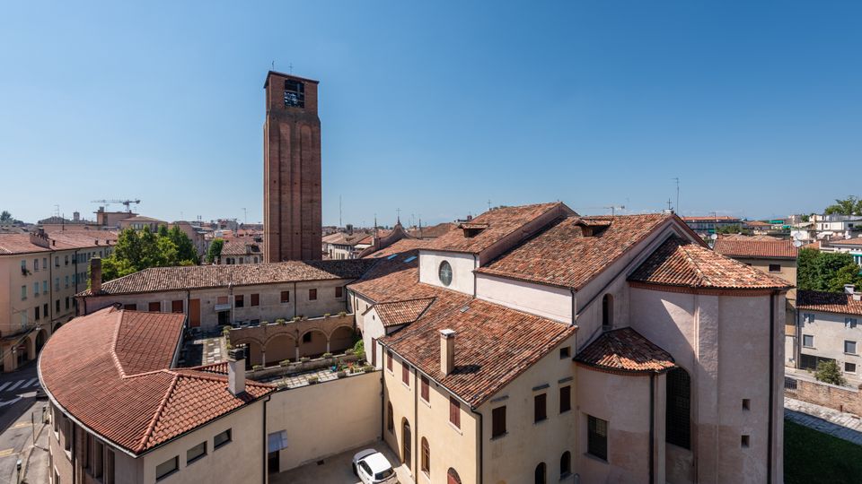 Veduta dall’alto di un complesso religioso con chiesa dai tetti in coppi rossi, cortile interno con porticato ad archi e un alto campanile in mattoni. Sullo sfondo si estende il tessuto urbano con edifici bassi e tetti in laterizio sotto cielo sereno.