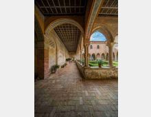 Chiostro storico in pietra con lungo porticato ad archi e soffitto a cassettoni, pavimentato in mattoni. A destra si apre un cortile interno con colonne, aiuole geometriche e vasi di piante, con facciate in muratura e cielo visibile tra gli archi.